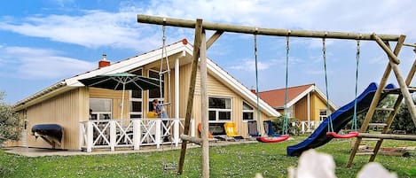 A playground with swings and a slide, a covered seating area, and a building with a sloped roof.