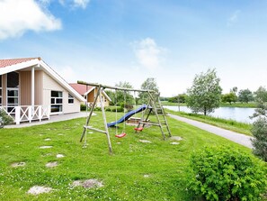 Ein Spielplatz mit Rutsche und Schaukeln, ein Haus mit rotem Dach und im Hintergrund ein Gewässer.