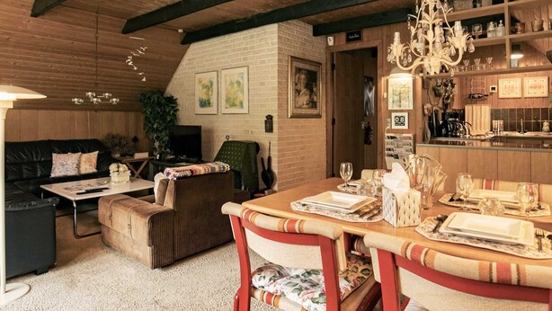 Ein gemütliches Wohnzimmer mit Holzbalkendecke, eine Essecke mit Tisch für vier Personen und eine durchgehende Tür sichtbare Küche.