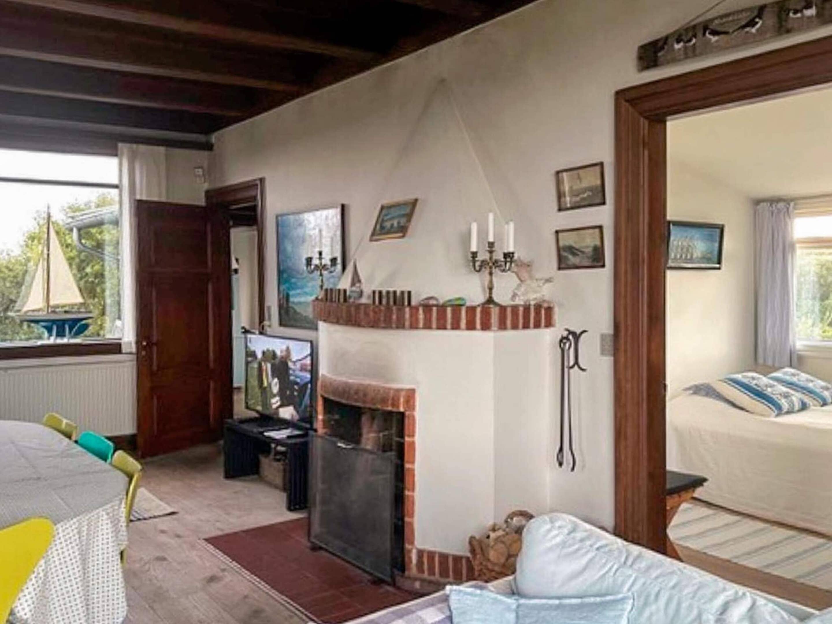 Une pièce avec une cheminée, un lit avec une literie à rayures, une fenêtre donnant sur un voilier, et une étagère murale agrémentée d’objets décoratifs.