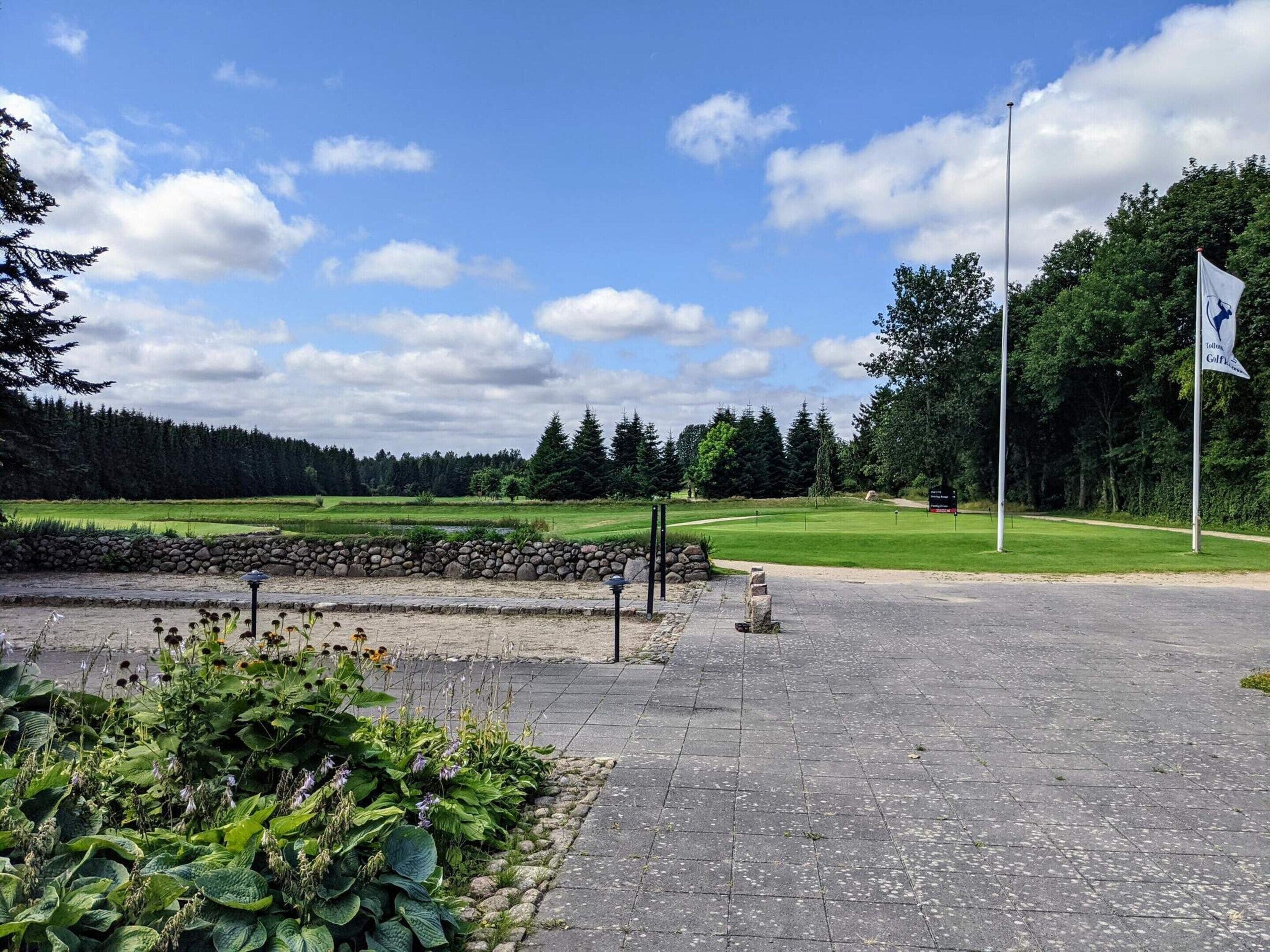 Un camino pavimentado que conduce a un campo de golf, con un muro de piedra y plantas a la izquierda.