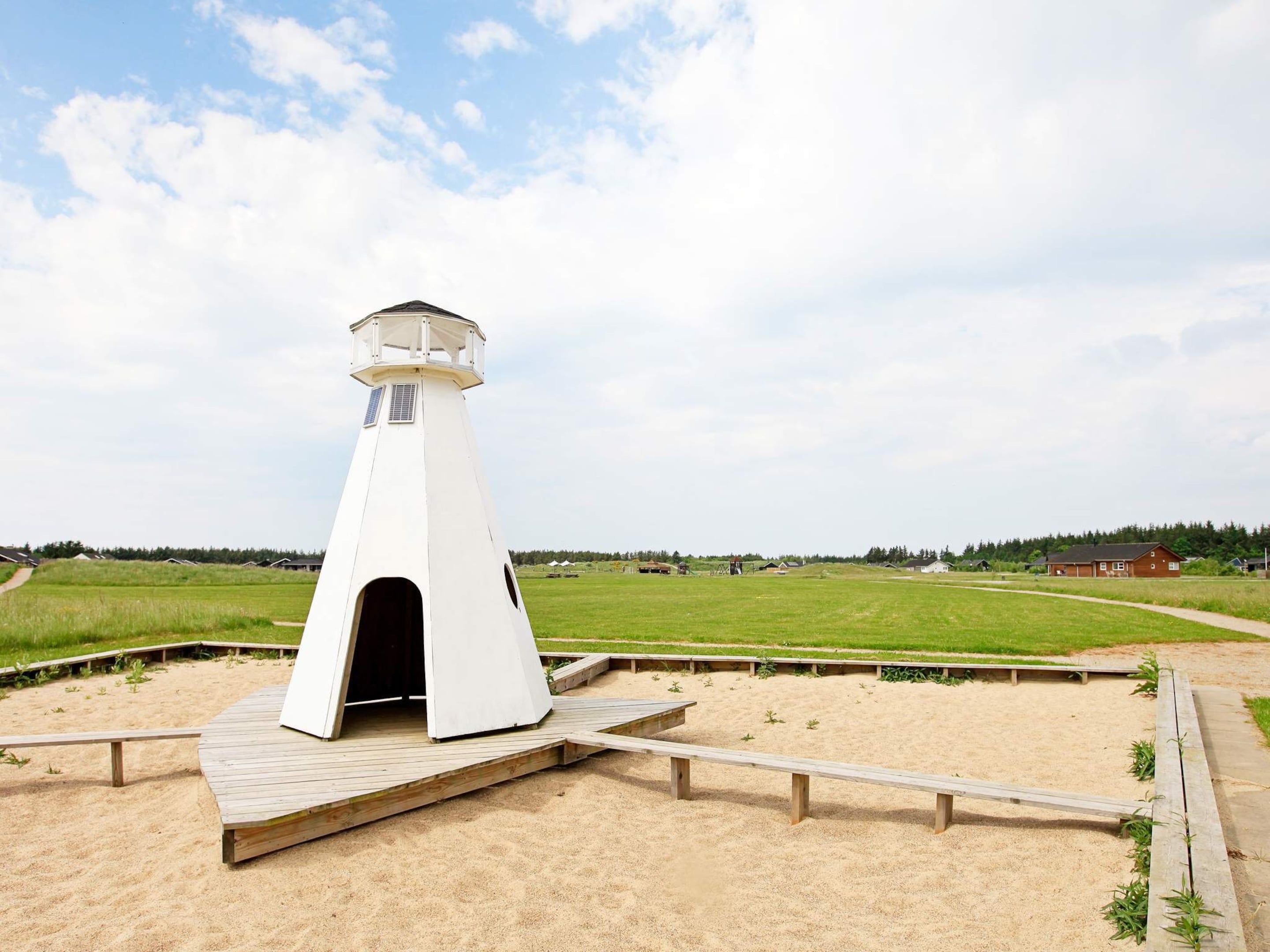 A white lighthouse structure with a wooden deck in front, surrounded by sand and grassy fields.