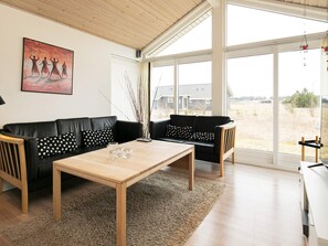 A modern living room with a wooden table, black leather sofas, and a large window offering a view of the outdoors.