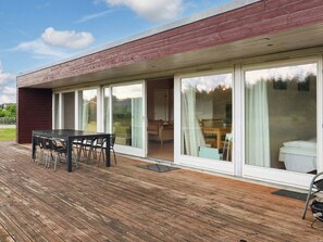 Une maison moderne dotée d’une terrasse en bois, d’un coin repas avec une table et des chaises, et de grandes portes-fenêtres coulissantes donnant sur un salon avec un canapé et une table basse.