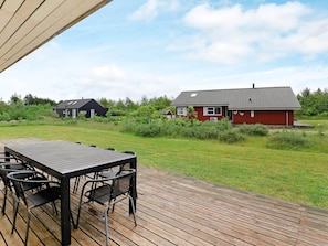 A wooden deck with a table and chairs, a red house with a tiled roof, and a grassy area with other houses in the background. - 6 Person Holiday Home in Brovst-by Traum (Brovst)
