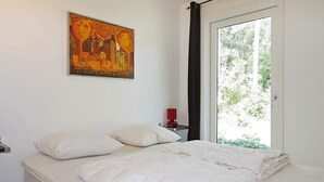 A bedroom with a bed, pillows, a painting, a nightstand, a red lamp, and a window with a view of greenery.