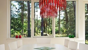A modern dining area with a white table and chairs, large windows with a forest view, and a red abstract hanging decoration.