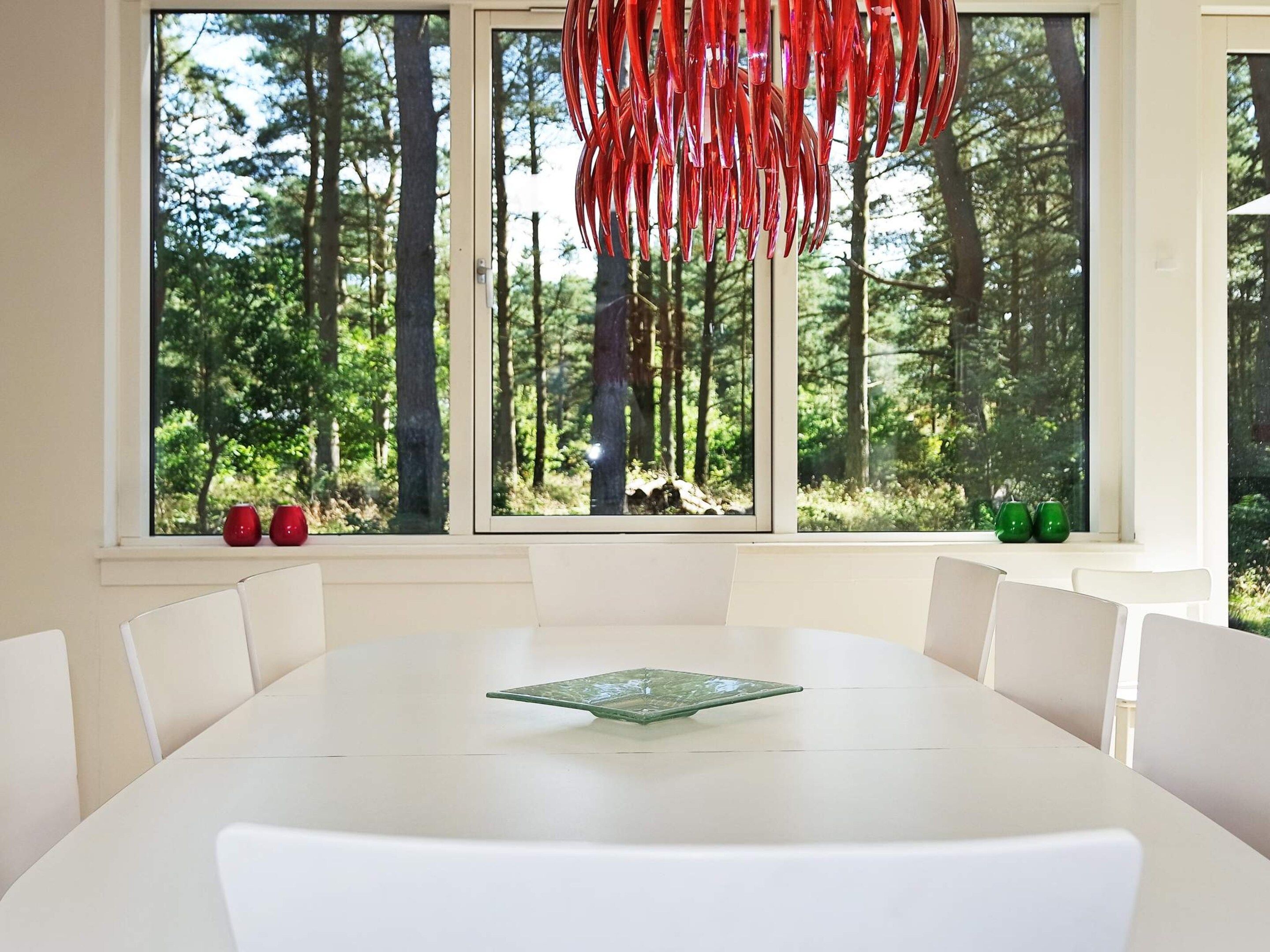 Un comedor con una mesa y sillas blancas, ventanas amplias con vistas al bosque y una decoración colgante roja.