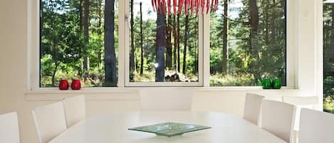 Un comedor con una mesa y sillas blancas, ventanas amplias con vistas al bosque y una decoración colgante roja.
