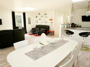 Una sala de estar moderna con un sofá negro, una mesa de comedor con sillas y una zona de cocina con un lavabo y armarios.