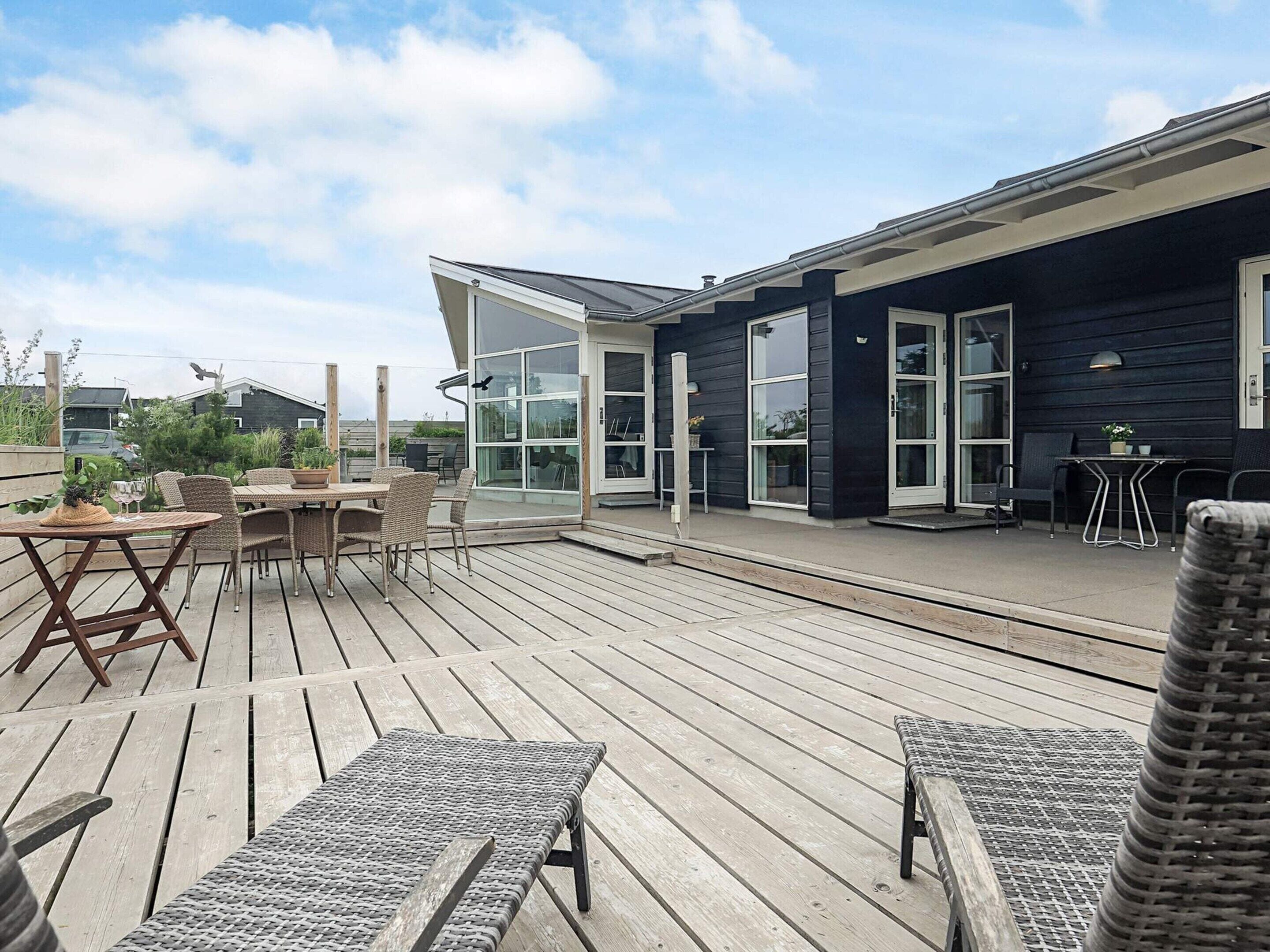 Une terrasse en bois, aménagée avec du mobilier d’extérieur, notamment une table et des chaises, et une porte vitrée donnant sur un espace abrité.