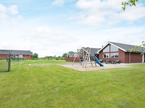 A playground with swings and a slide, surrounded by a grassy area and a building with large windows. - 5 Star Holiday Home in Nordborg (Nordborg)
