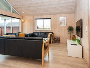 A modern living room with a black leather sofa, wooden flooring, and a large window offering a view of the outdoors. - 5 Star Holiday Home in Nordborg (Nordborg)