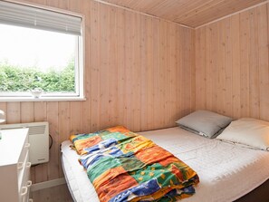 Ein Schlafzimmer mit einer holzgetäfelten Wand, einem Bett mit bunter Decke, einem Kopfkissen und einem Fenster mit Jalousien.