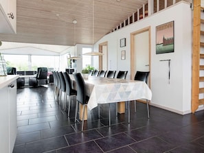 Un espace repas avec une longue table, des chaises noires et une nappe blanche. Le plafond est orné de poutres en bois et une grande fenêtre laisse entrer la lumière naturelle.