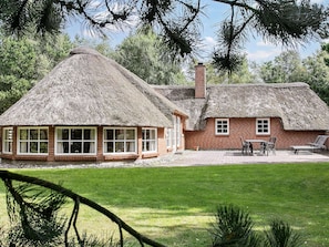 A thatched-roof house with a brick extension, a patio with chairs, and a lawn.