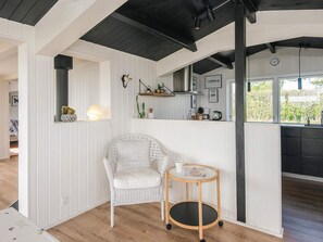 A room with a white wicker chair, a small round table, a window with a view of greenery, and a kitchen area with a black countertop. - 4 Star Holiday Home in Ebeltoft (Ebeltoft)