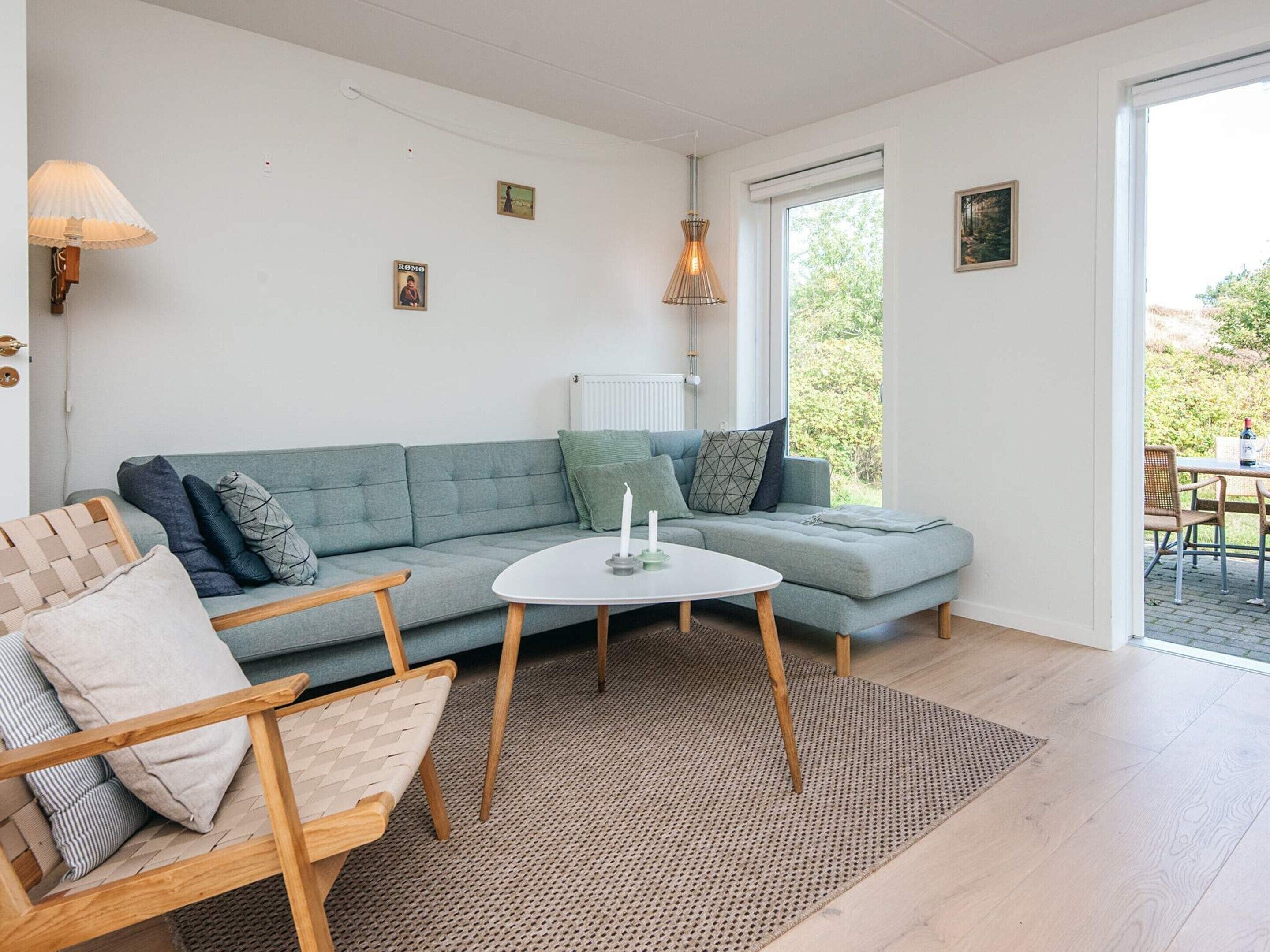 A modern living room with a grey sectional sofa, a small round table, and a woven chair. There is a window with a view of greenery outside.