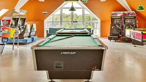 Une table de billard dans une pièce aux murs orange, avec des jeux d’arcade et une grande fenêtre.