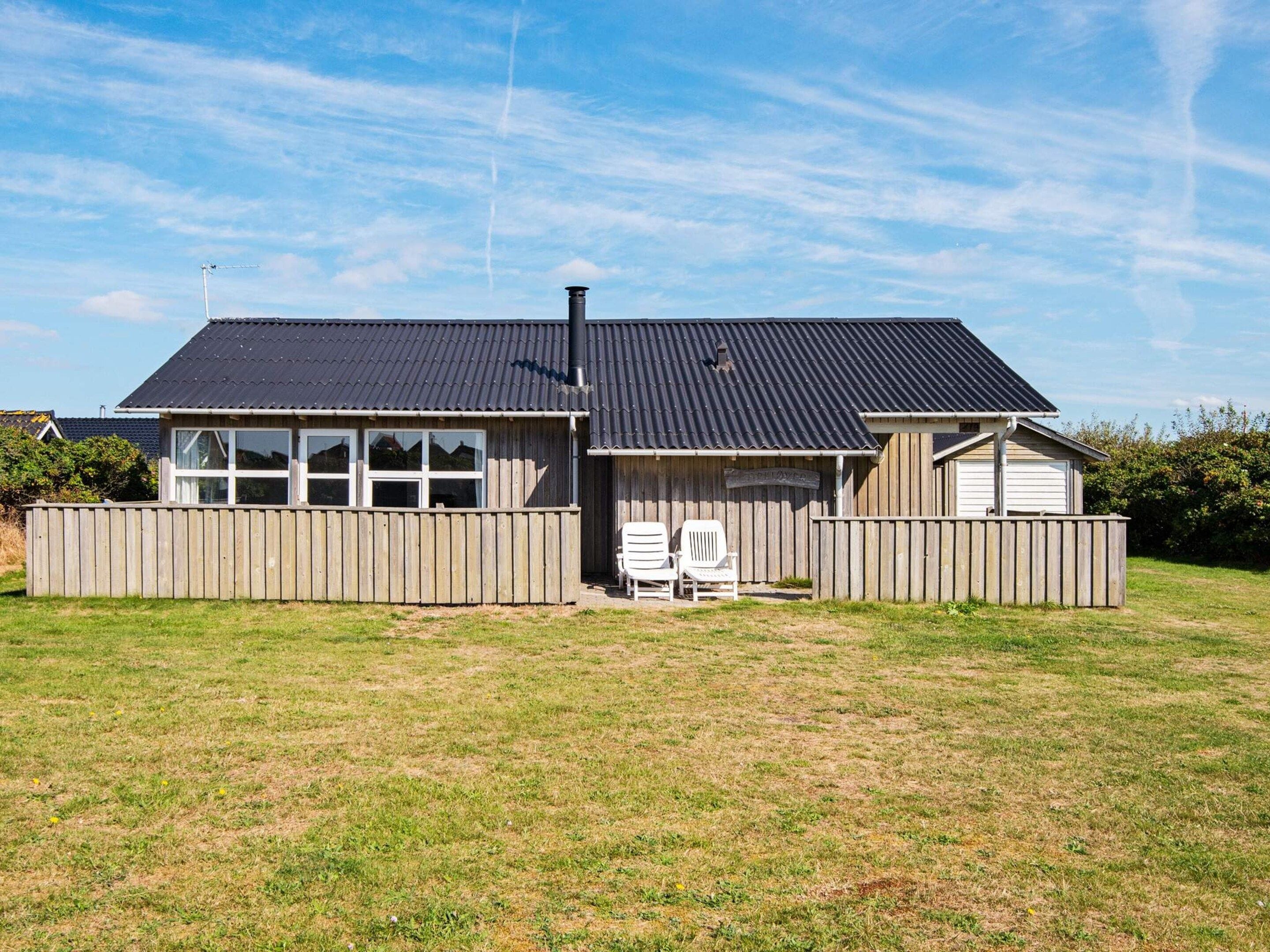 A single-story house with a dark roof, a fenced yard, and two white chairs outside.
