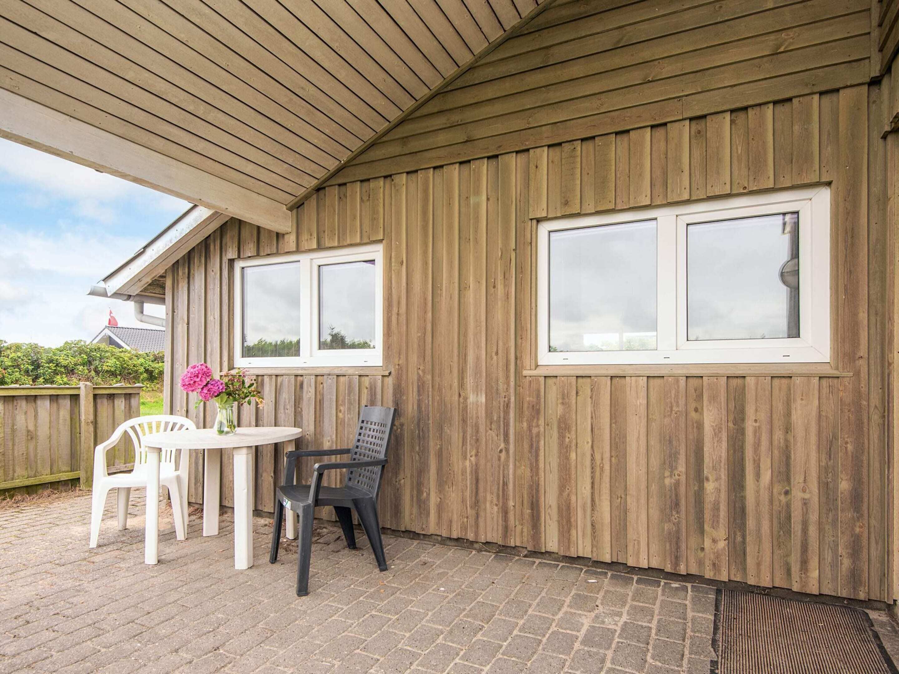 A covered patio area with a wooden wall, two windows, a table with chairs, and a flower vase.