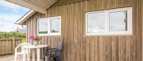 A covered patio area with a wooden wall, two windows, a table with chairs, and a flower vase.