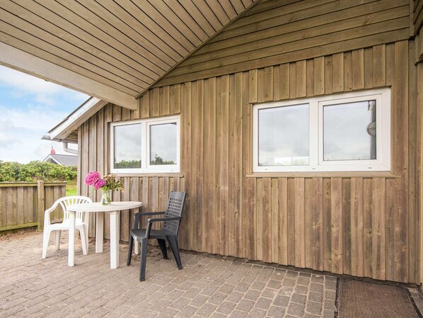 A covered patio area with a wooden wall, two windows, a table with chairs, and a flower vase.