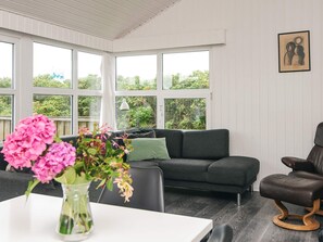 A cozy living room with a dark grey sofa, a brown leather chair, and a white table with a vase of pink flowers. Large windows offer a view of greenery outside.