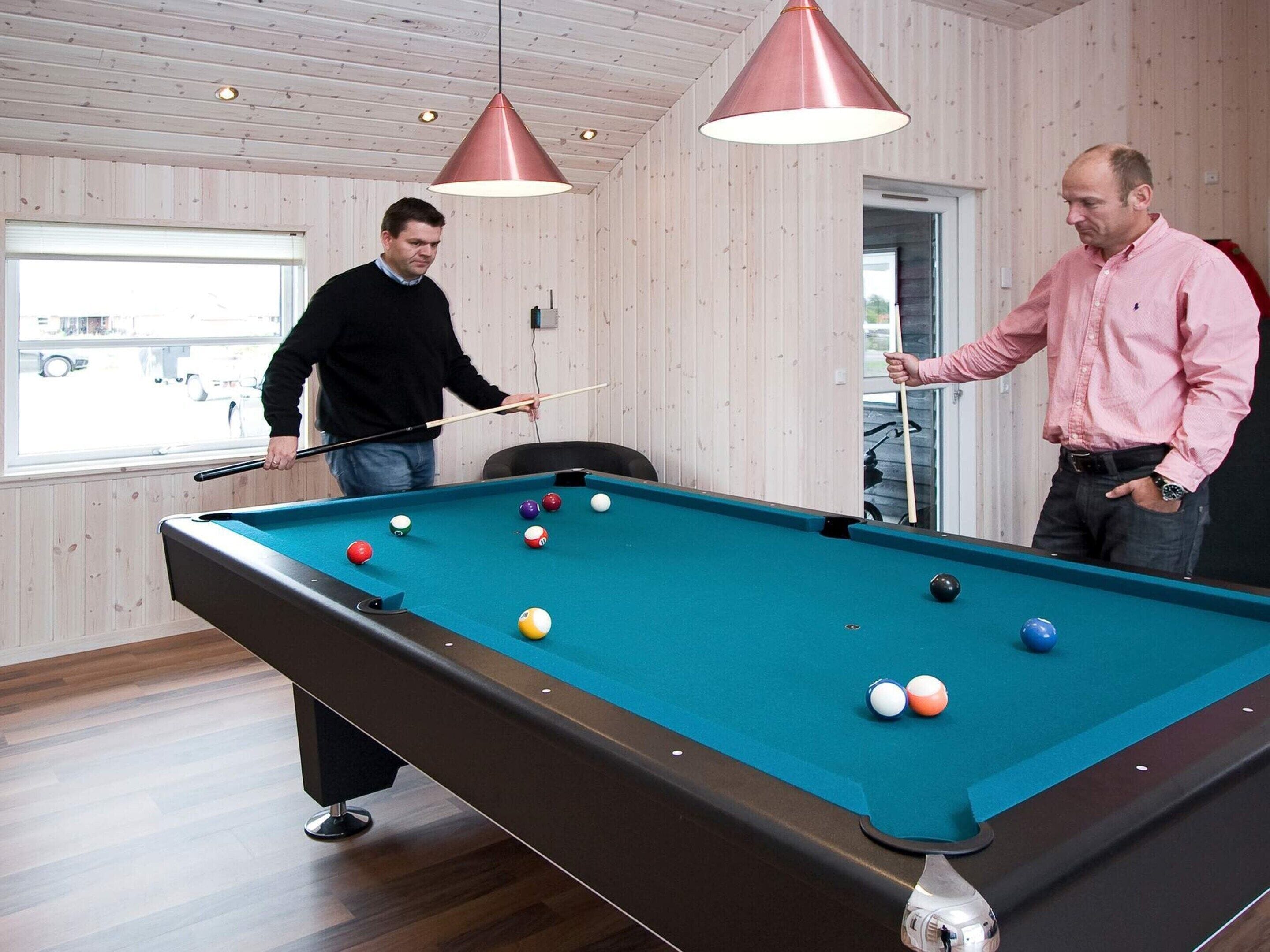 A pool table with balls, two people playing, wooden walls, and pendant lights.