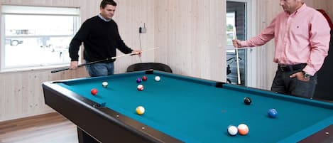 A pool table with balls, two people playing, wooden walls, and pendant lights.