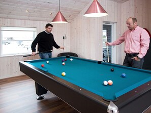 Ein Billardtisch mit Kugeln, zwei Personen beim Spielen, Holzwände und Pendelleuchten.
