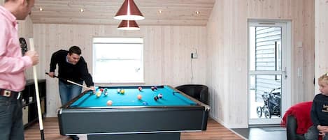 Una mesa de billar en una habitación con paredes de madera y techo inclinado, una ventana con luz natural y una puerta que da al exterior.
