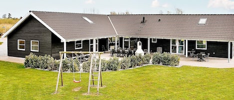 A black building with a sloped roof, a covered patio area with outdoor seating, and a grassy lawn with a swing set.