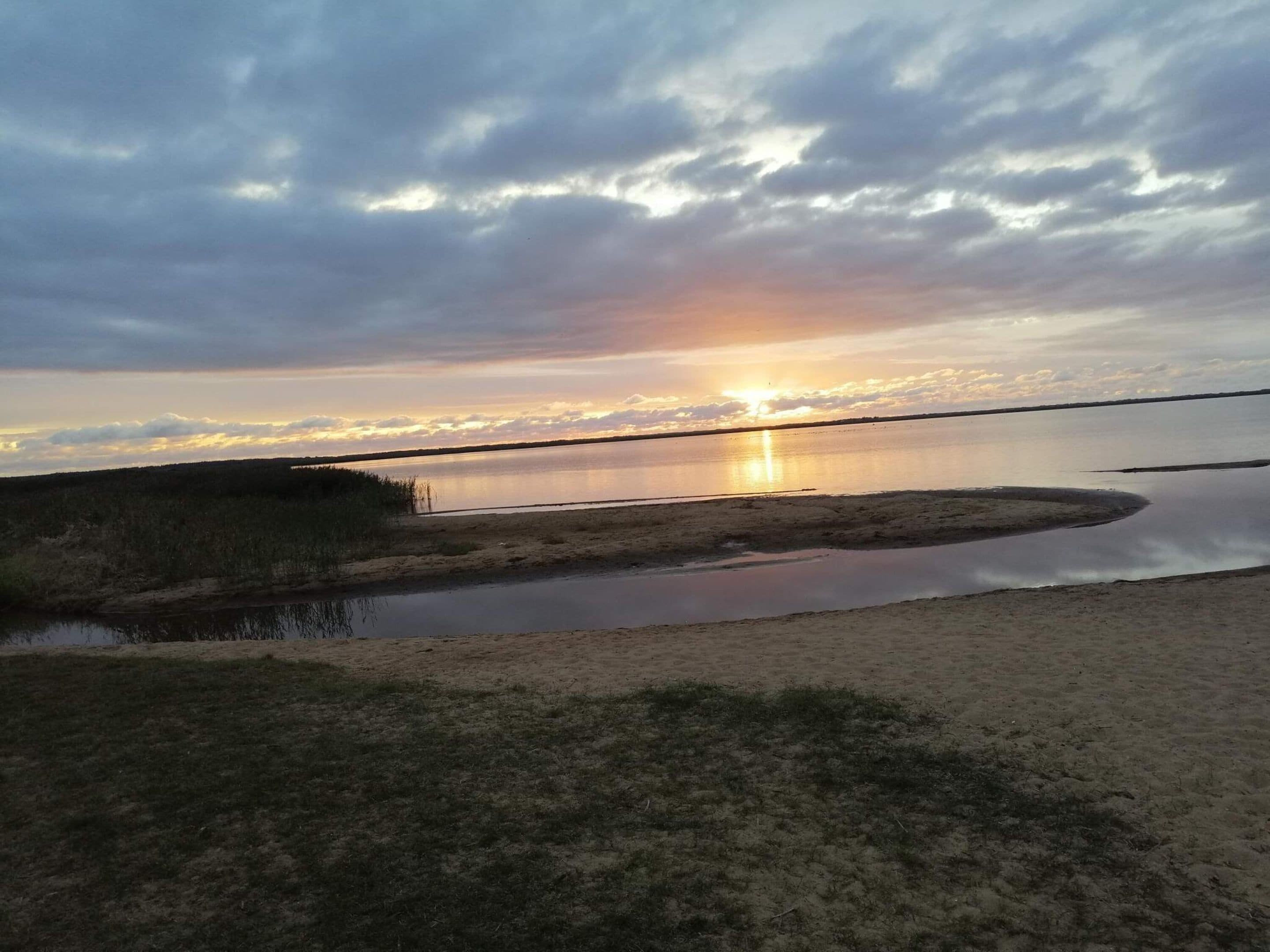 Ett naturligt landskap med en vattenyta som speglar himlen, omgivet av gräsbevuxna områden och en avlägsen horisont.