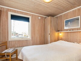 Un dormitorio con paneles de madera, una cama con colcha blanca, una silla, una ventana con vista a edificios y un estante con un cuadro.
