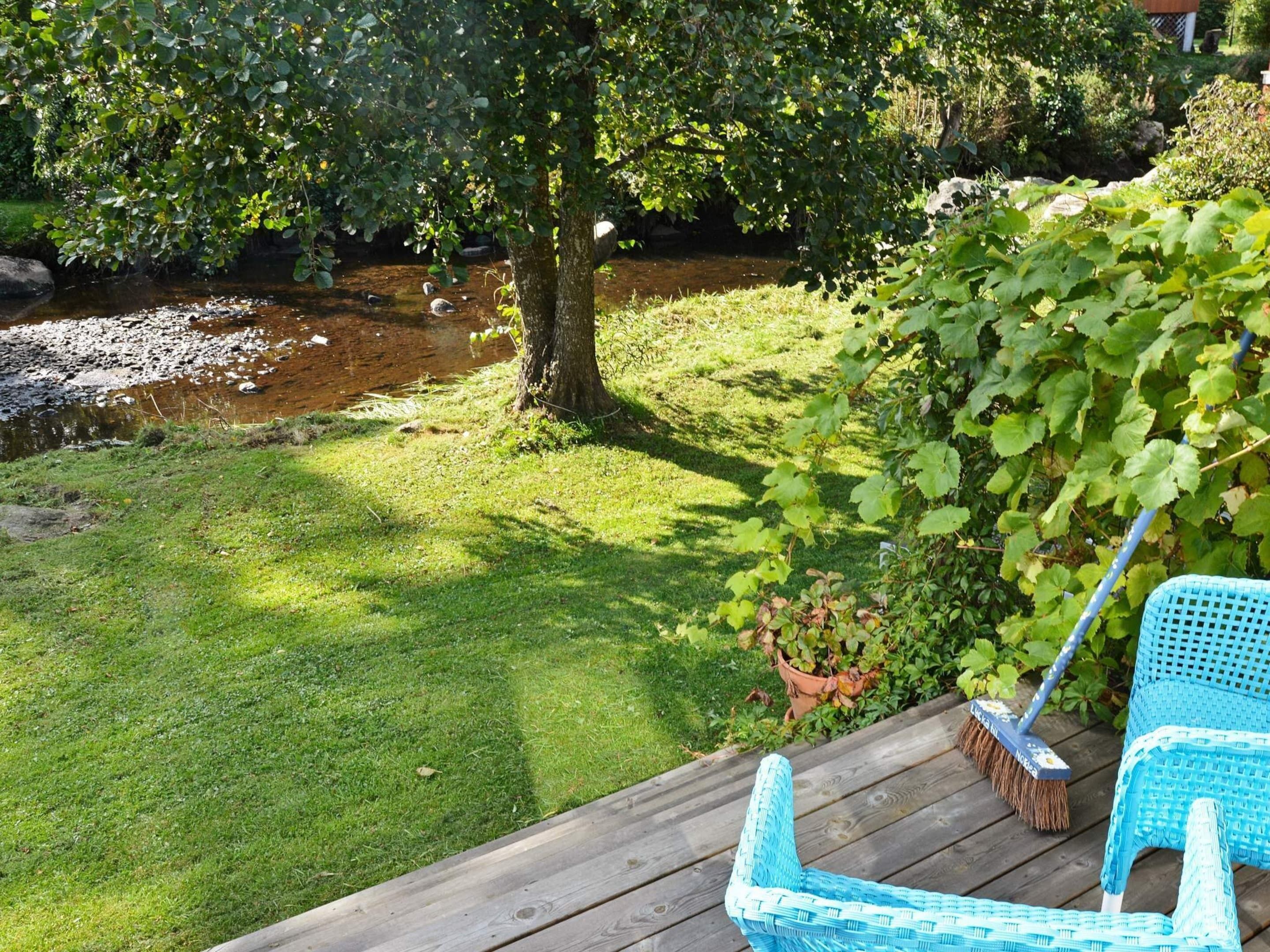 Una terrazza in legno con una sedia blu e una scopa, affacciata su una zona erbosa con un albero e un piccolo ruscello.