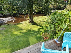 A wooden deck with a blue chair and a broom, overlooking a grassy area with a tree and a small stream.