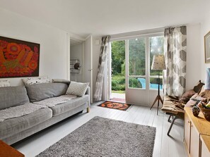 A living room with a grey sofa, a wooden cabinet, and a large window with curtains. - Holiday Home in Sölvesborg (Sölvesborg)