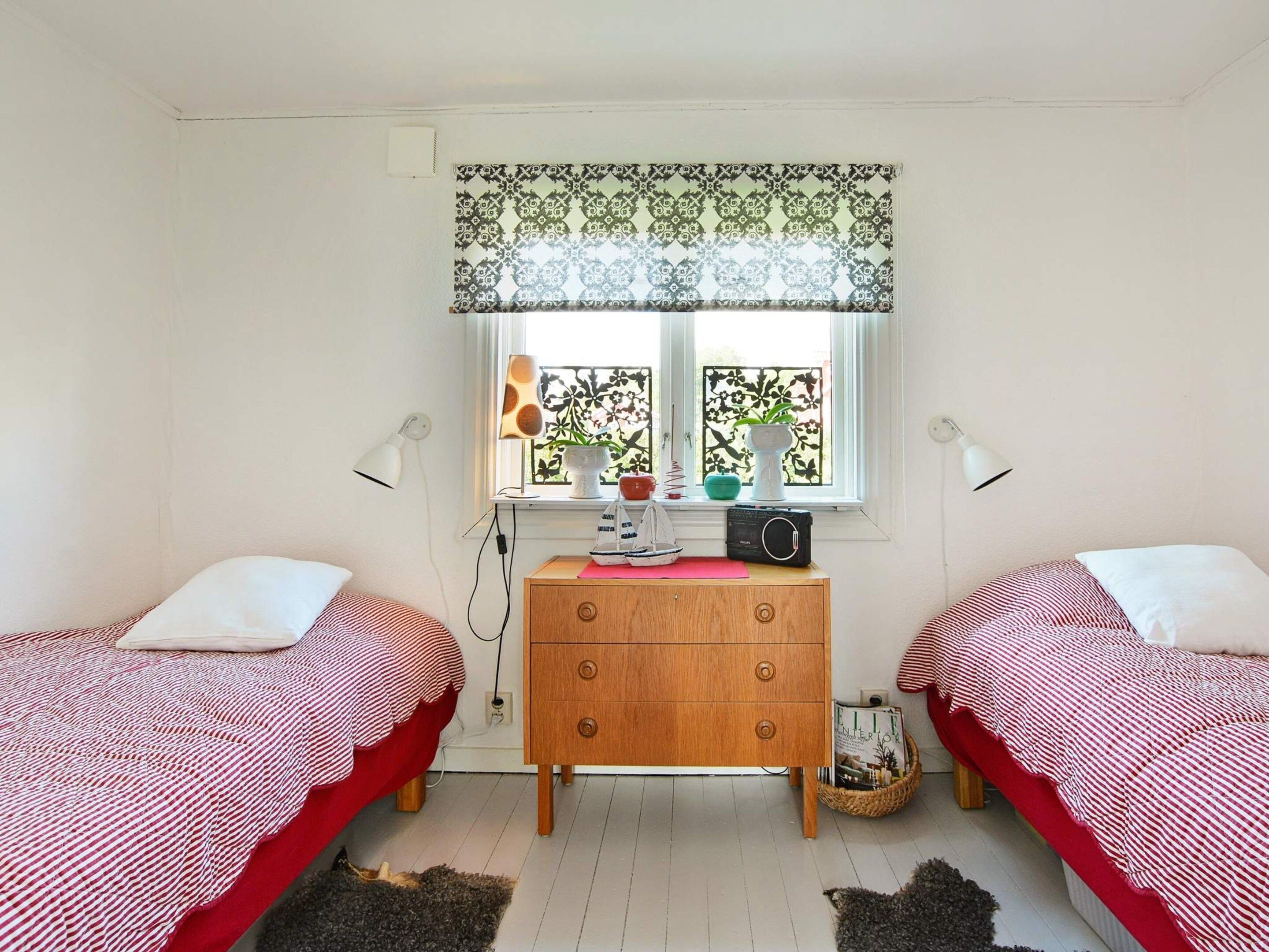 A bedroom with two single beds, a wooden dresser, a window with patterned curtains, and a small table with a radio and decorative items.