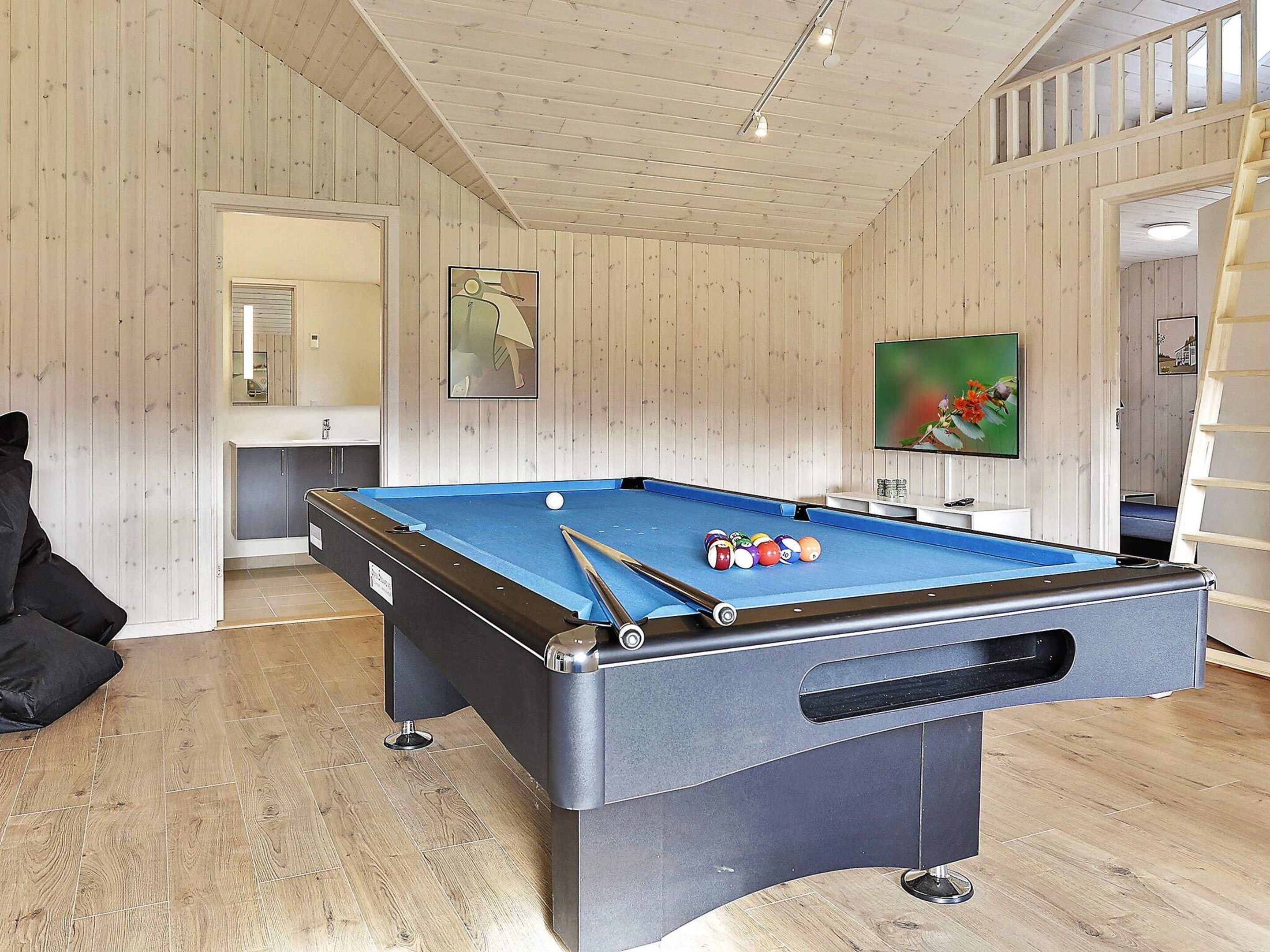 A pool table with balls and cues, a television, and a ladder in a room with wooden walls and a tiled floor.