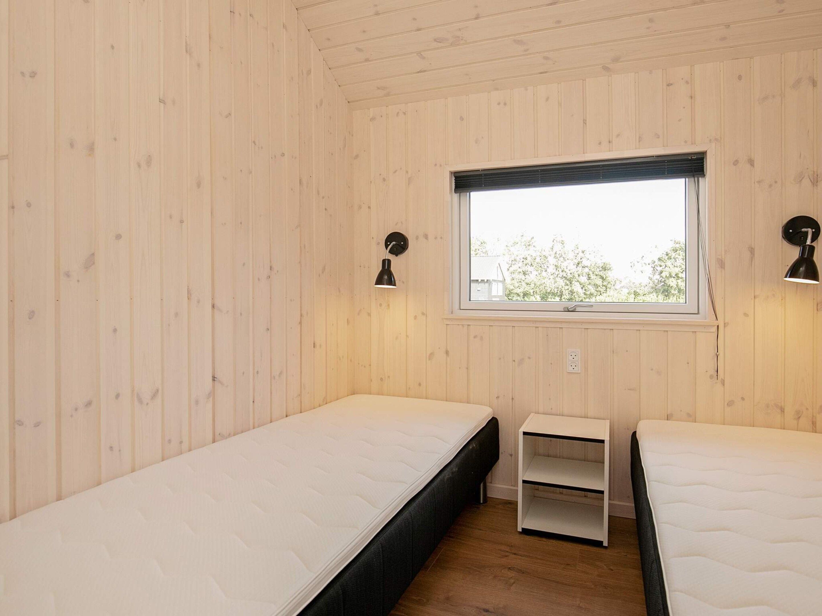 A room with two single beds, a window with blinds, a wall-mounted lamp, and a small white nightstand.
