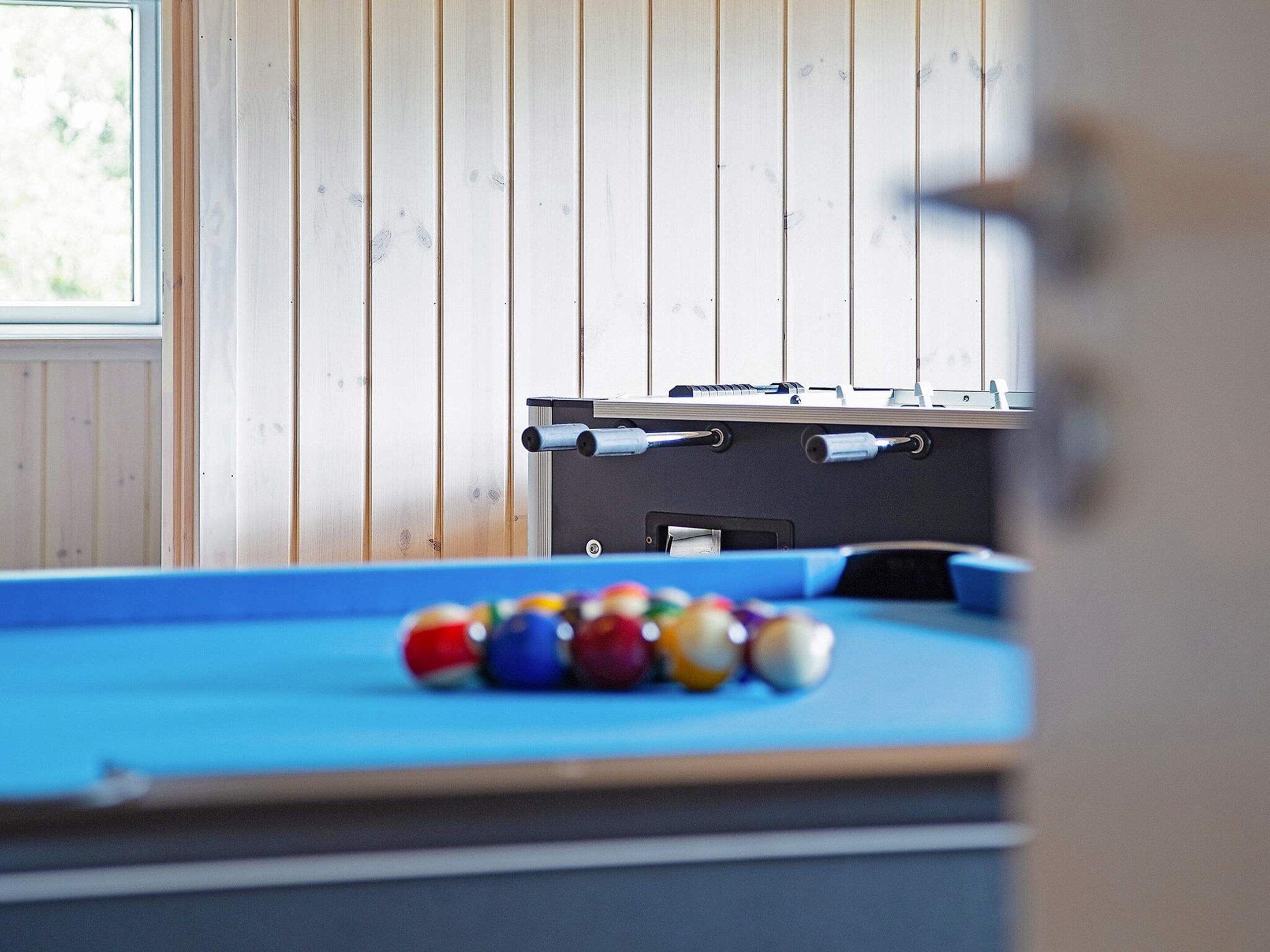 A pool table with balls in the foreground, a pool table in the background, and a window with a view of greenery.