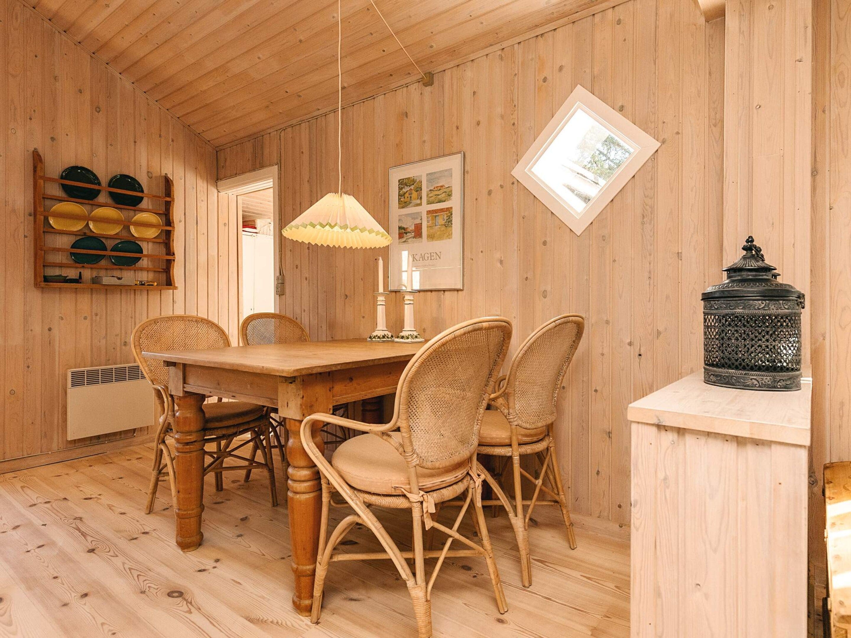 Een kamer met houten lambrisering, een eettafel, rieten stoelen, een hanglamp, een lijst met een afbeelding en een decoratieve lantaarn.
