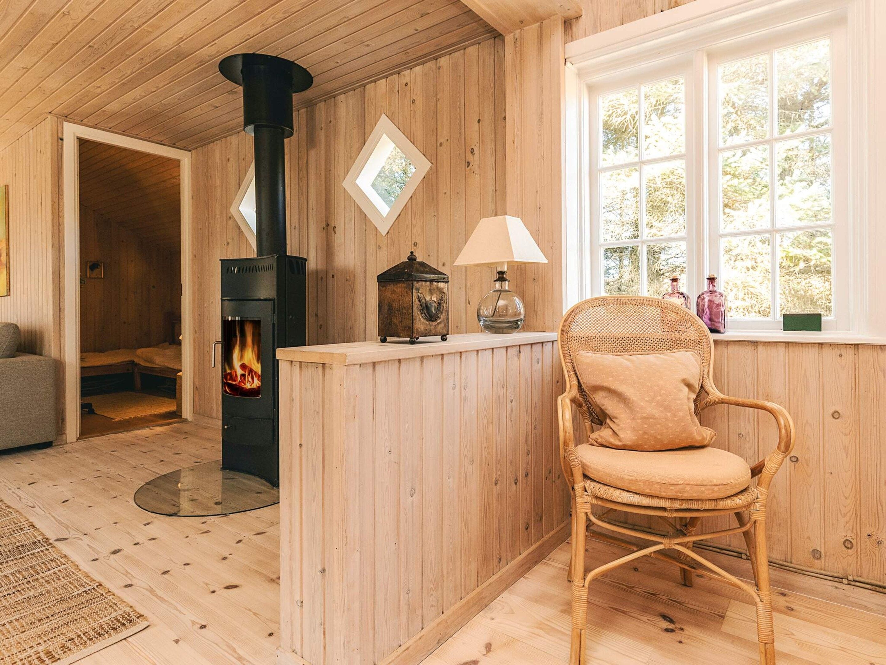 Ein gemütliches Zimmer mit Holzvertäfelung, einem Kamin, einem Korbsessel und einem Fenster mit Blick auf Bäume.