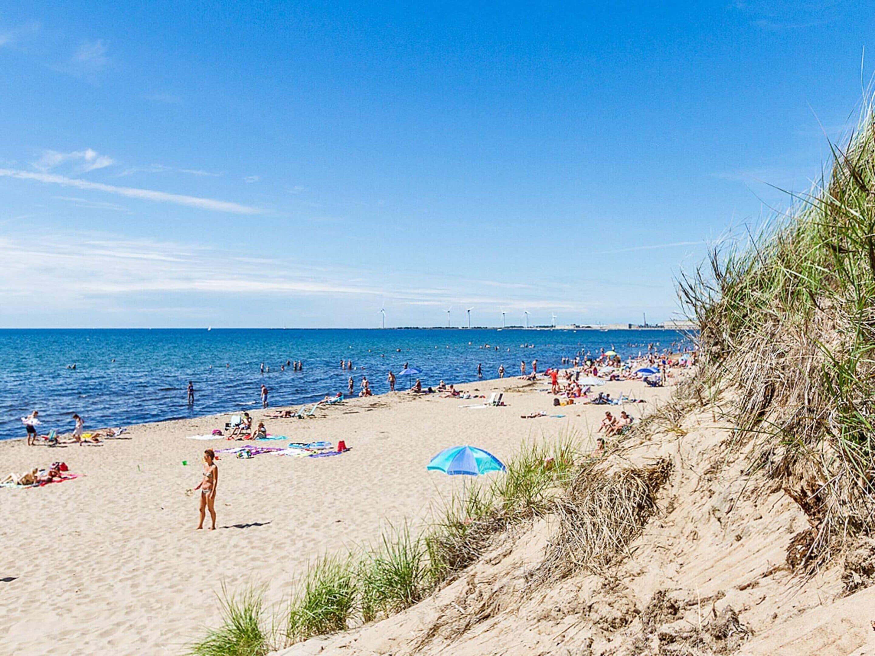 En strand med folk som solar och badar, en klar himmel och en vindkraftverk i fjärran.