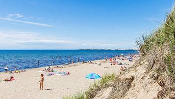 En strand med folk som solar och badar, en klar himmel och en vindkraftverk i fjärran.