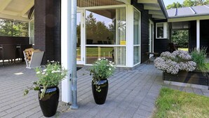 A covered patio area with a brick floor, two potted plants, a metal pipe, and a glass door leading to an outdoor seating area. - 6 Person Holiday Home in Hojslev-by Traum (Hojslev)