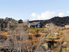 Una casa con techo oscuro y varias ventanas, rodeada por un paisaje natural con árboles dispersos y una colina rocosa al fondo.