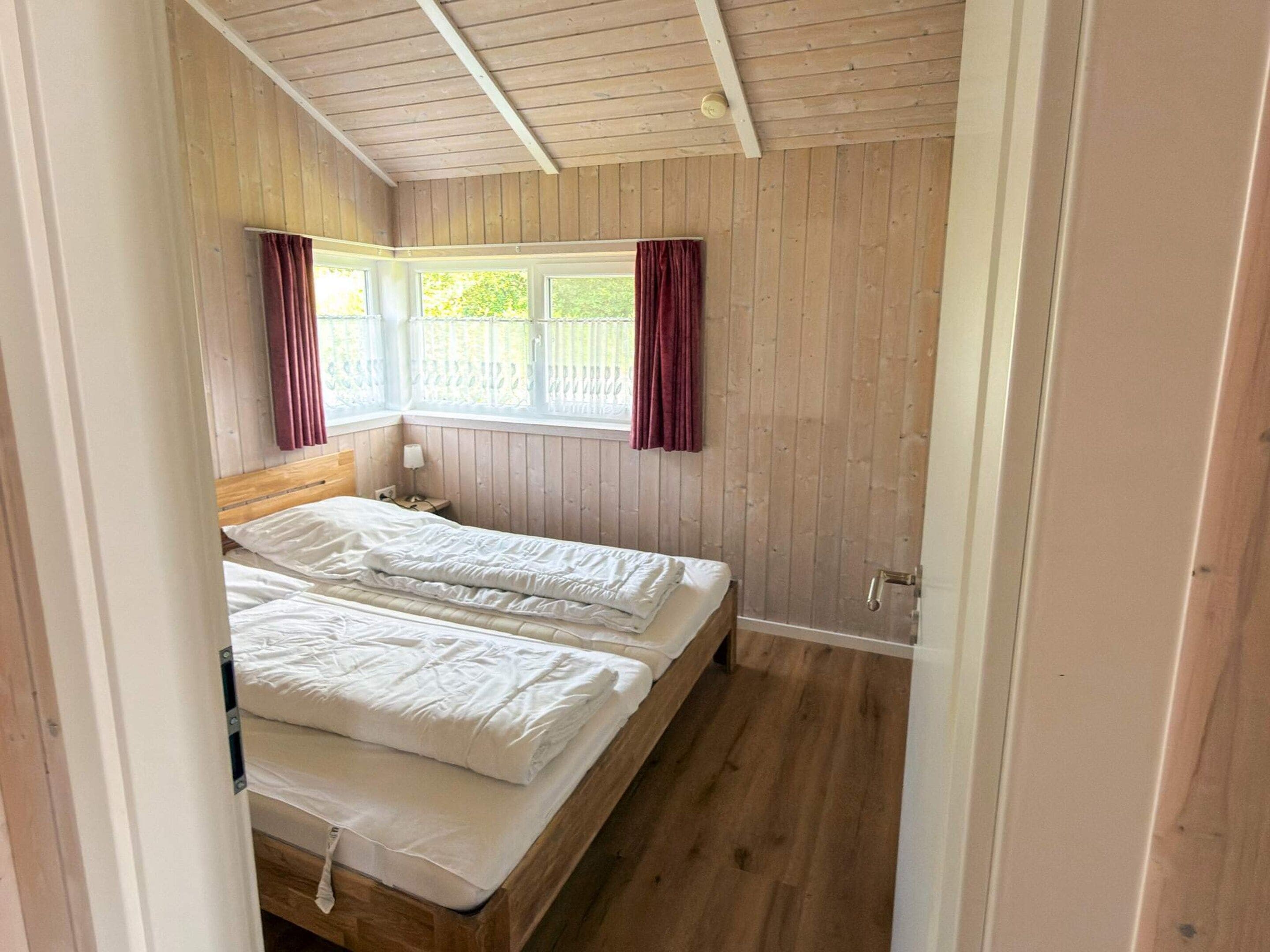 A bedroom with a wooden ceiling, a bed with white bedding, a window with curtains, and a nightstand with a lamp.