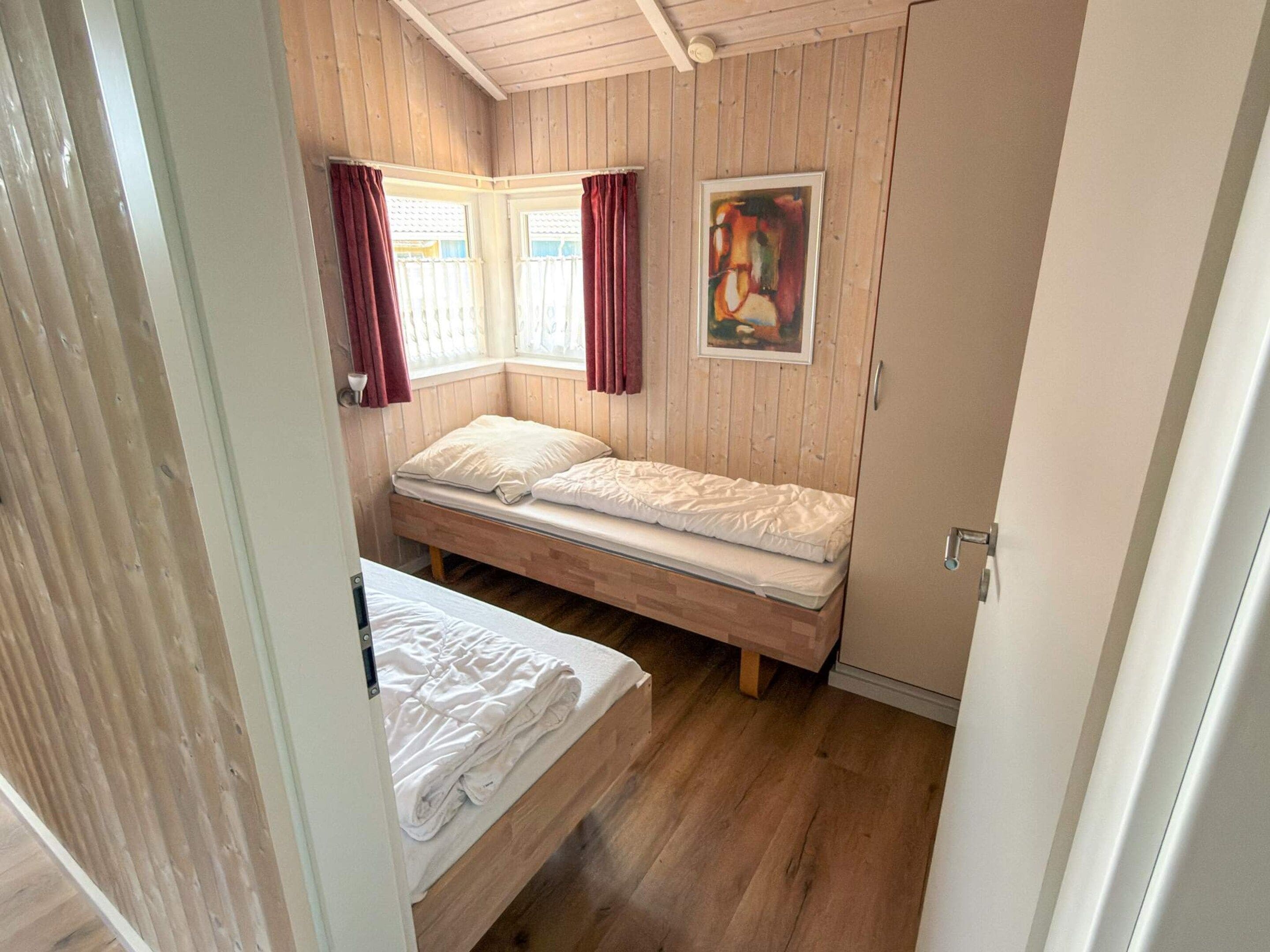 Et lille, enkelt værelse med trægulv, to senge, et vindue med gardiner og et billede på væggen.